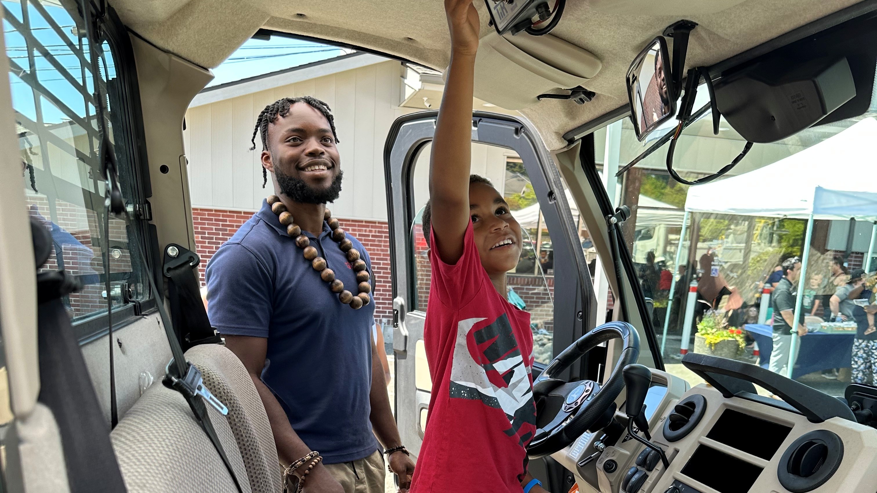 A young boy sits in a truck and plays with the controls while his father looks on.