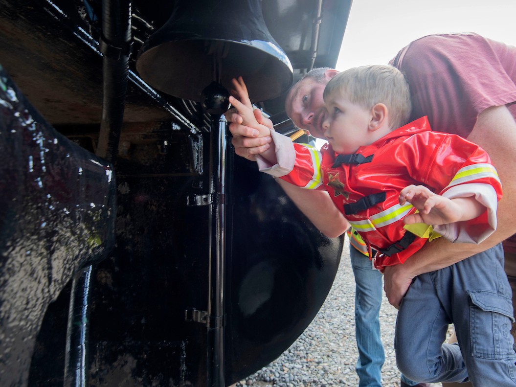 A toddler boy is held up by an adult man to touch part of a vehicle. 