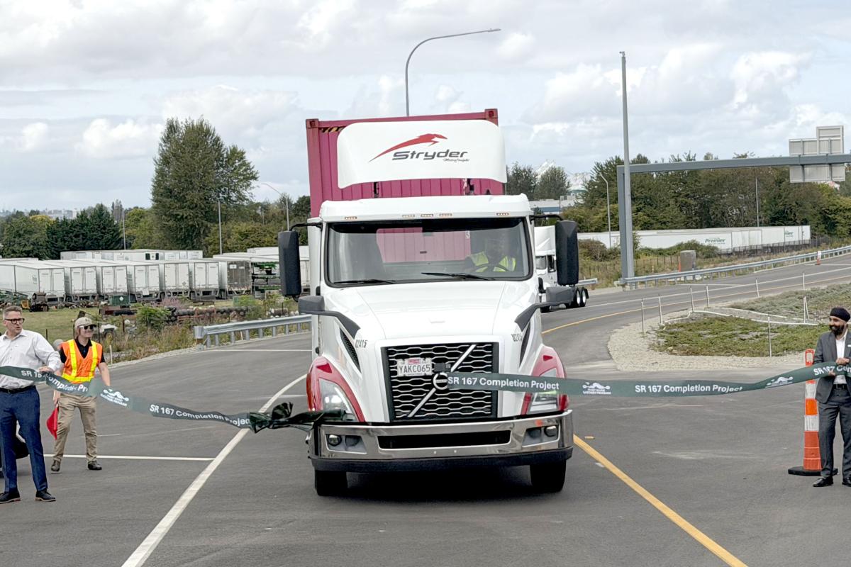 A semi truck drives through a ribbon to celebrate the opening of SR167 expressway as community partners hold the ribbon.